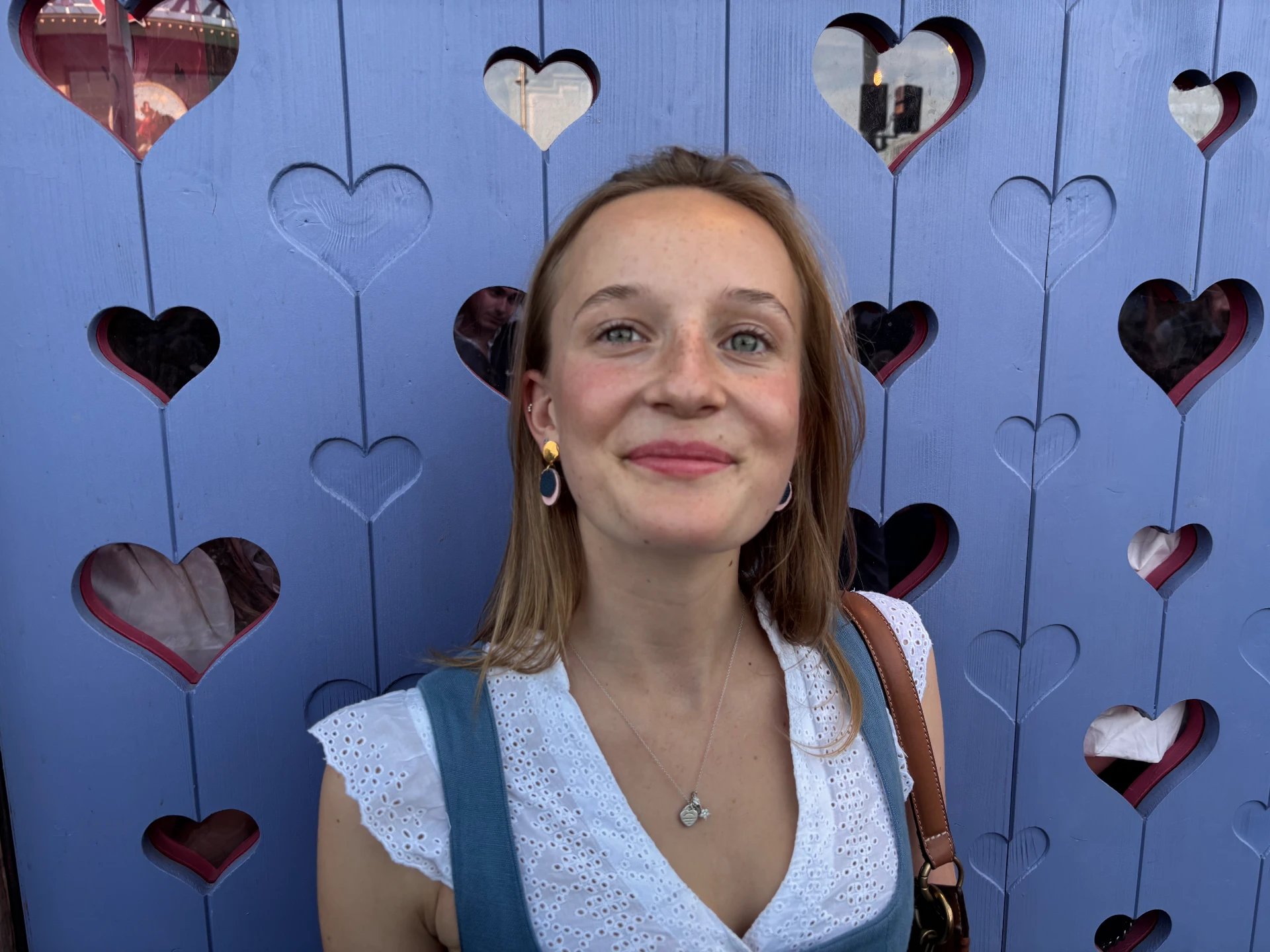 Smiling woman wearing Cleo leather earrings in front of a blue wooden wall with heart cutouts